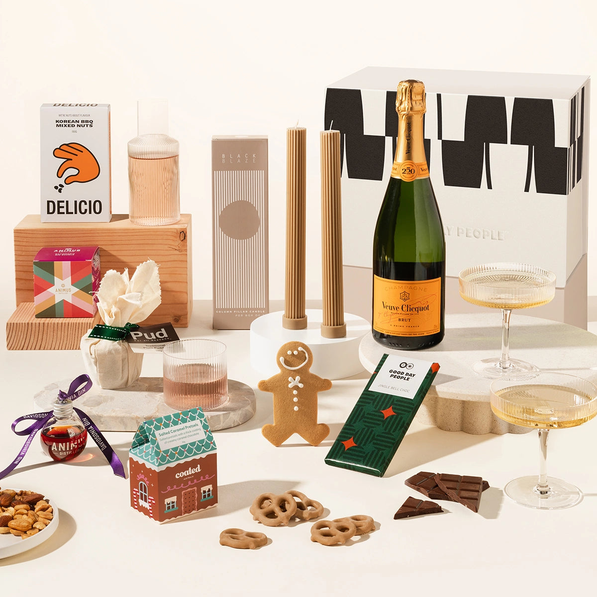 Assorted holiday gift hamper including a bottle of champagne, delicio korean bbq nuts, pillar candles, ferm living carafe and glass set, 2 champagne coupes, chocolate, gingerbread man, salted caramel pretzels and a gin bauble. All packaged in a beautiful gift box.