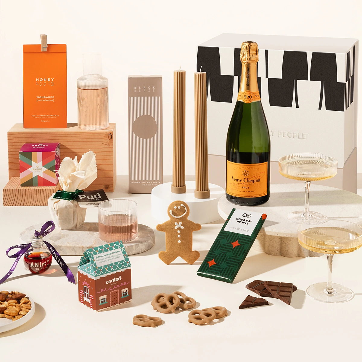 Assorted holiday gift hamper including a bottle of champagne, delicio korean bbq nuts, pillar candles, ferm living carafe and glass set, 2 champagne coupes, chocolate, gingerbread man, salted caramel pretzels and a gin bauble. All packaged in a beautiful gift box.