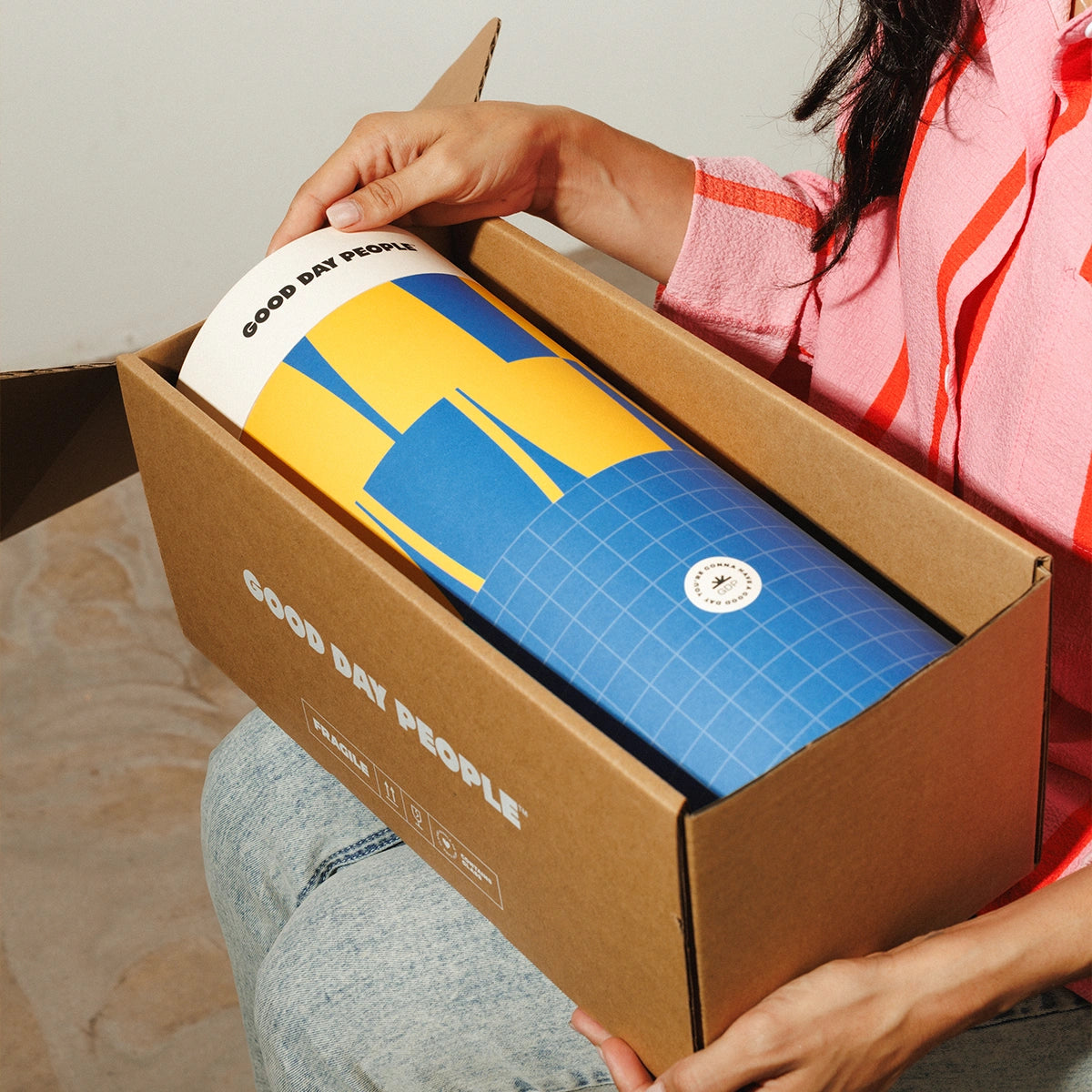 A person holding an open cardboard box while revealing a rolled Good Day People product wrapped in brown paper. The product has a blue and yellow geometric design with a white top section and visible Good Day People branding.