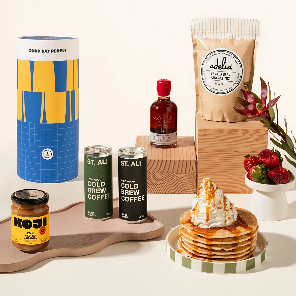 A styled product arrangement on a warm cream background featuring a blue and yellow cylinder, two ST. ALI cold brew coffee cans, a jar of Koji caramel spread, pancake mix, maple syrup, a stack of pancakes topped with whipped cream and caramel sauce, and a small white cup of strawberries, all arranged on wooden blocks and a wooden board.
