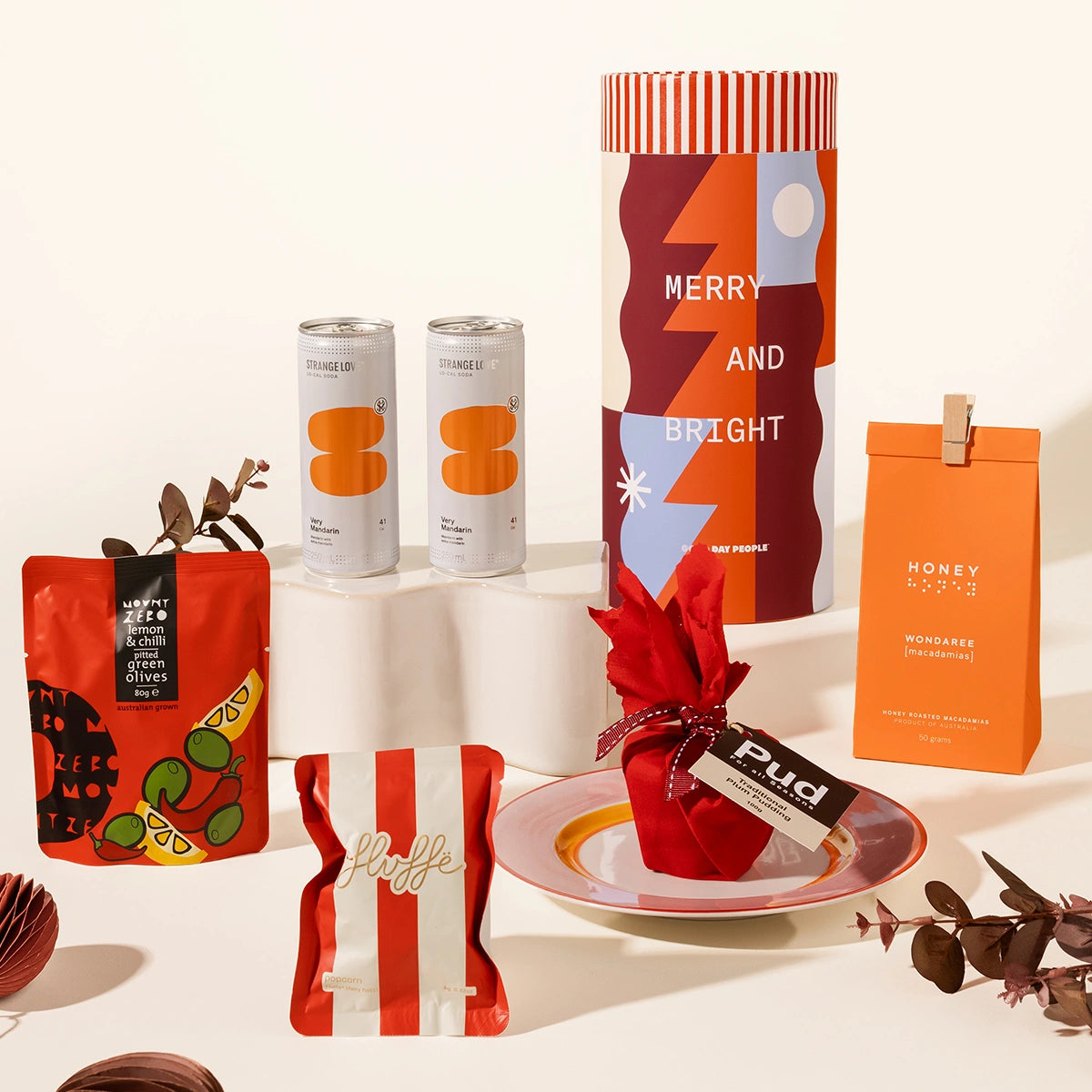 A curated gift assortment featuring various packaged food products, including a large cylindrical gift packaging canister with a Christmas design, macadamias, fairy floss, 2 x strangelove sodas, mount zero olives and a mini christmas pudding.