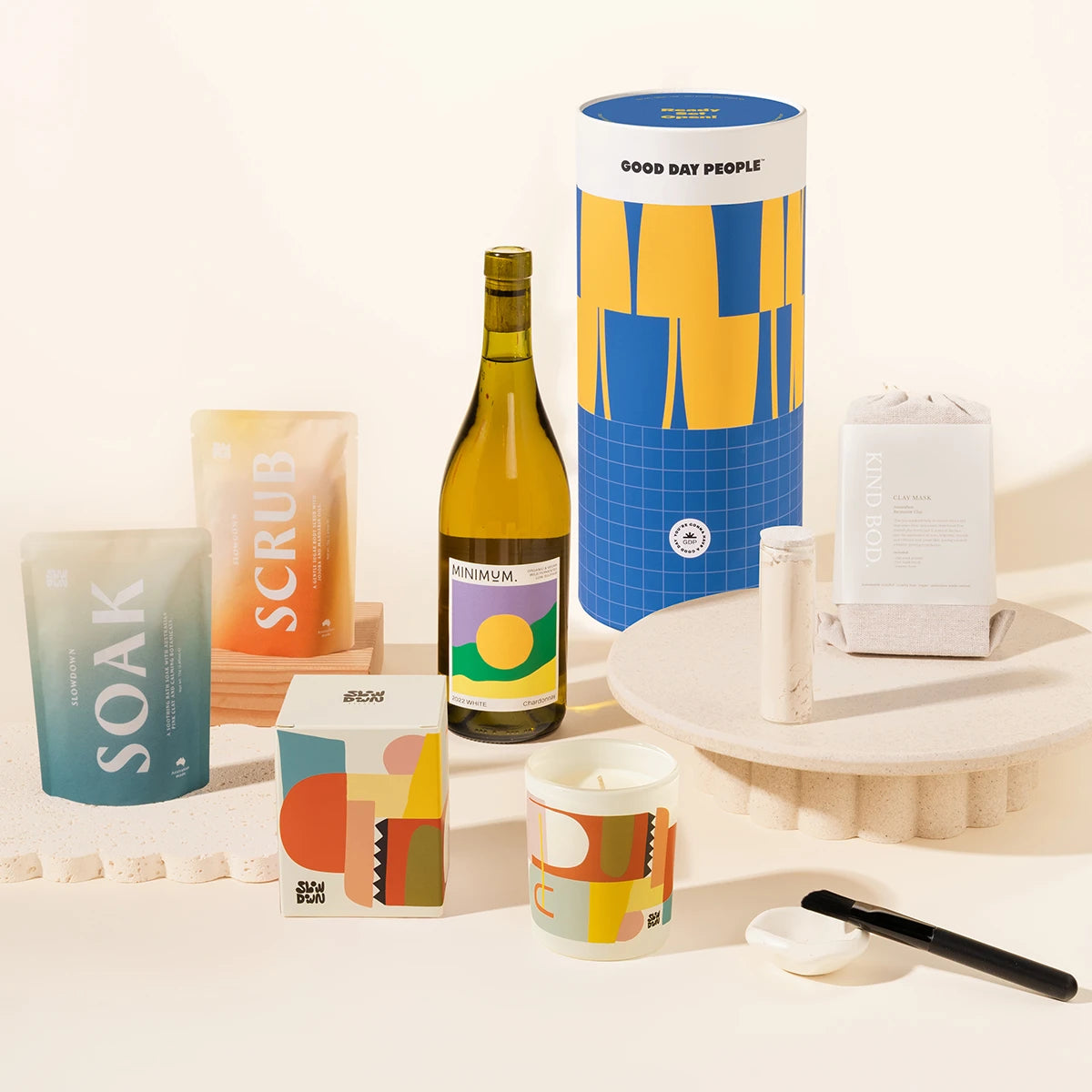 A curated self-care gift set arranged on a warm off-white surface and pedestal. Visible items include a tall blue-and-yellow cylinder GOOD DAY PEOPLE, a wine bottle, a blue gradient pouch labeled SOAK, a peach pouch labeled SCRUB, a face mask set with brush and ceramic holder, a candle in a white jar with abstract multicolor artwork.
