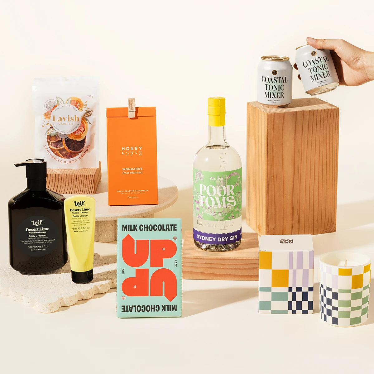 A curated gin gift set featuring Poor Tom's gin, strangelove tonics, Leif, orange garnish, arranged on wooden blocks and a white surface. The set includes a milk chocolate bar, macadamias, candle.