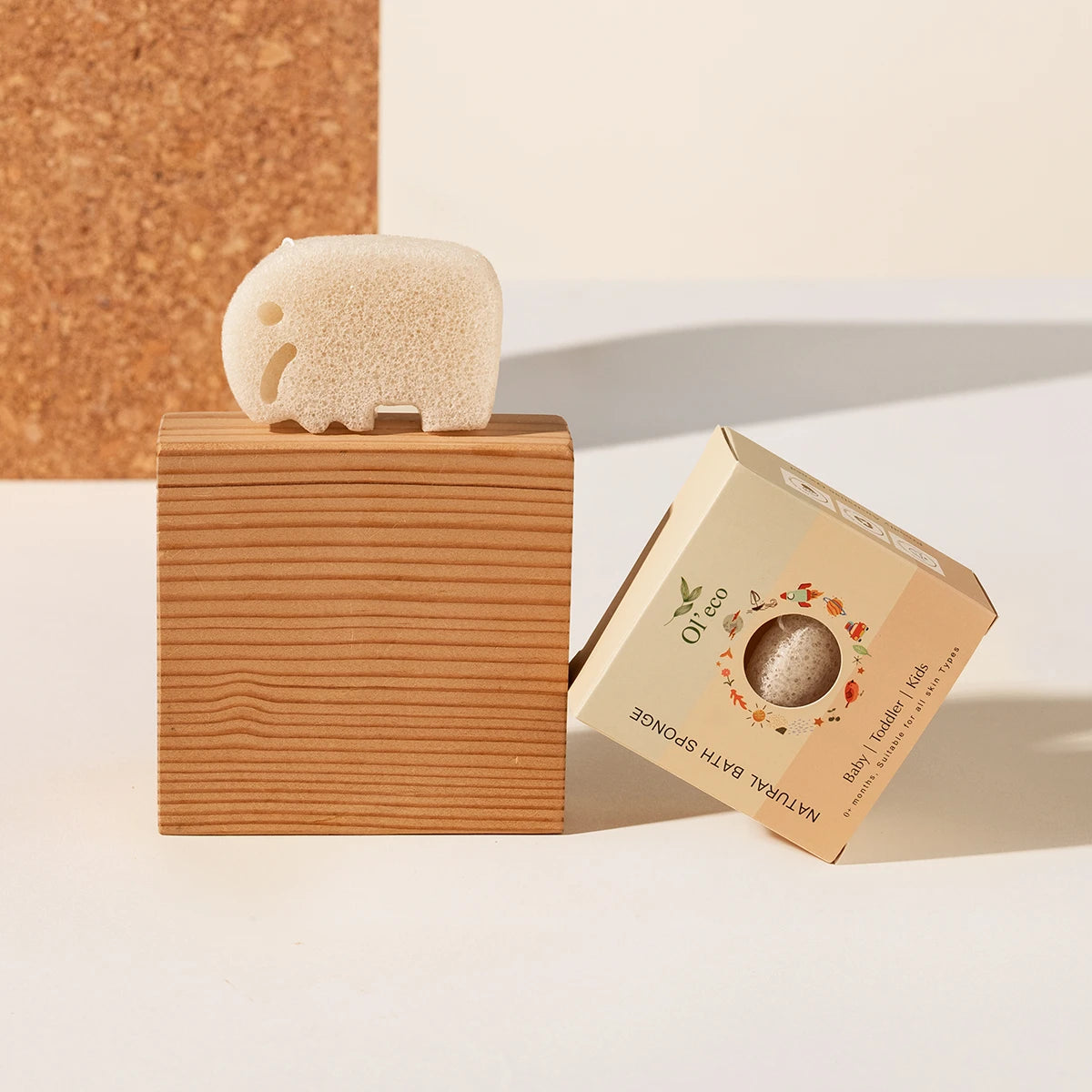 A konjac beige elephant-shaped bath sponge displayed on a wooden block next to its kraft paper packaging box, set against a clean white and cork background.