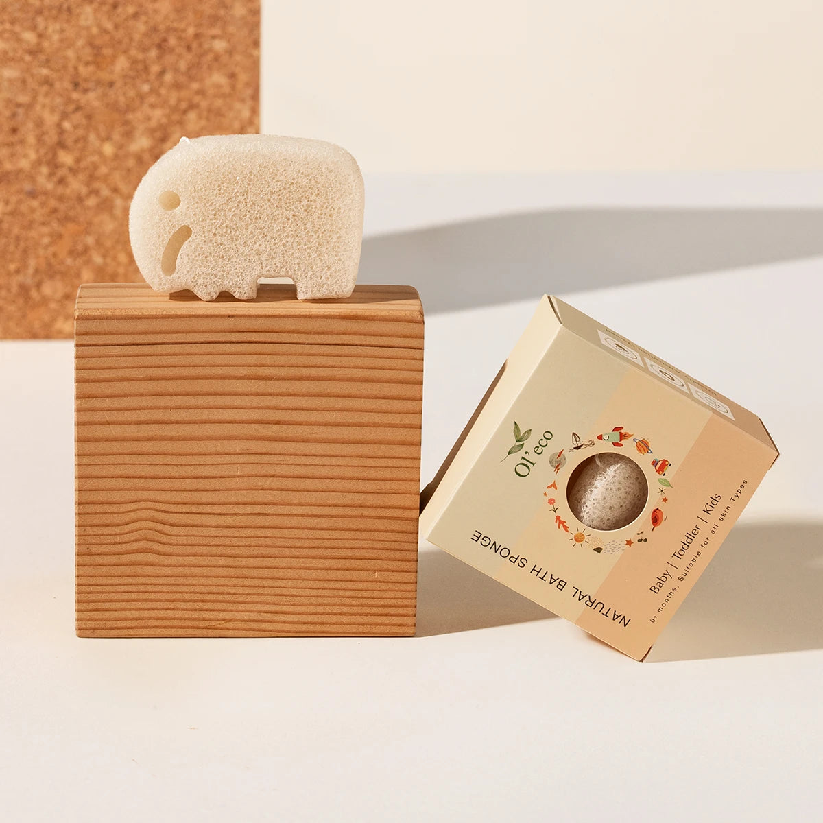 A natural beige konjac bath sponge shaped like an elephant resting on a wooden block, next to its kraft paper packaging box labeled.
