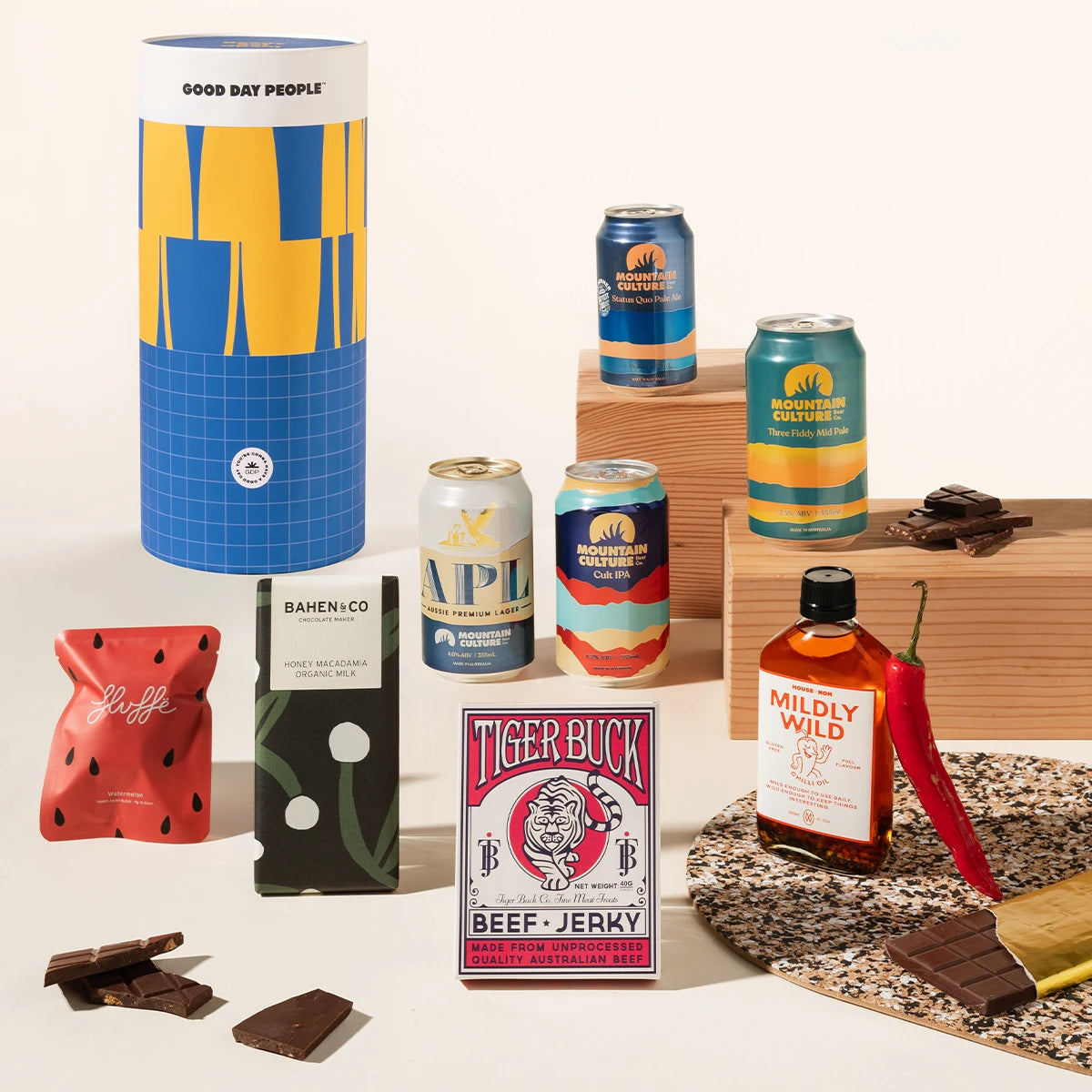 A curated gift assortment featuring a large blue and yellow Good Day People gift canister, 4 craft beer cans, a red watermelon-flavored fairy floss pouch, a chocolate bar package, a Tiger Buck beef jerky box, and a bottle of Mildly Wild chilli oil and chocolate pieces arranged around it on a clean light beige studio background.