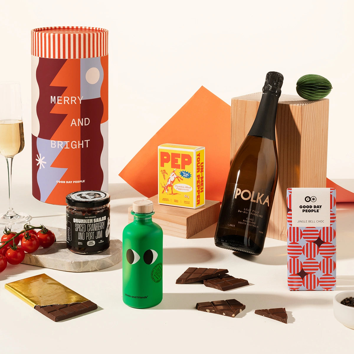 A foodie Christmas gift hamper styled on a cream backdrop. The packaging features a festive design with geometric shapes in red and blue on a canister. A glass bottle of non-alc sparkling, alongside a bold yellow box of gourmet pepper, a Christmassy block of chocolate, a green bottle of olive oil and a jar of jam.