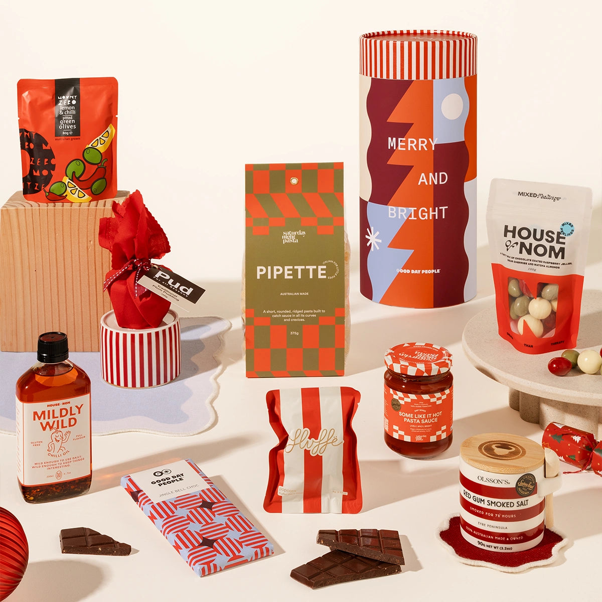 A curated gift assortment of packaged snacks, chocolates, and a bottle of spirits arranged on a white studio background. Visible items include a cylindrical tube reading “MERRY AND BRIGHT,” a green-and-orange checkered “PIPETTE” box, a white bag labeled “HOUSE OF NOM,” a bottle labeled “MILDLY WILD,” wrapped chocolates, chocolate bars, candy, and small gift boxes.