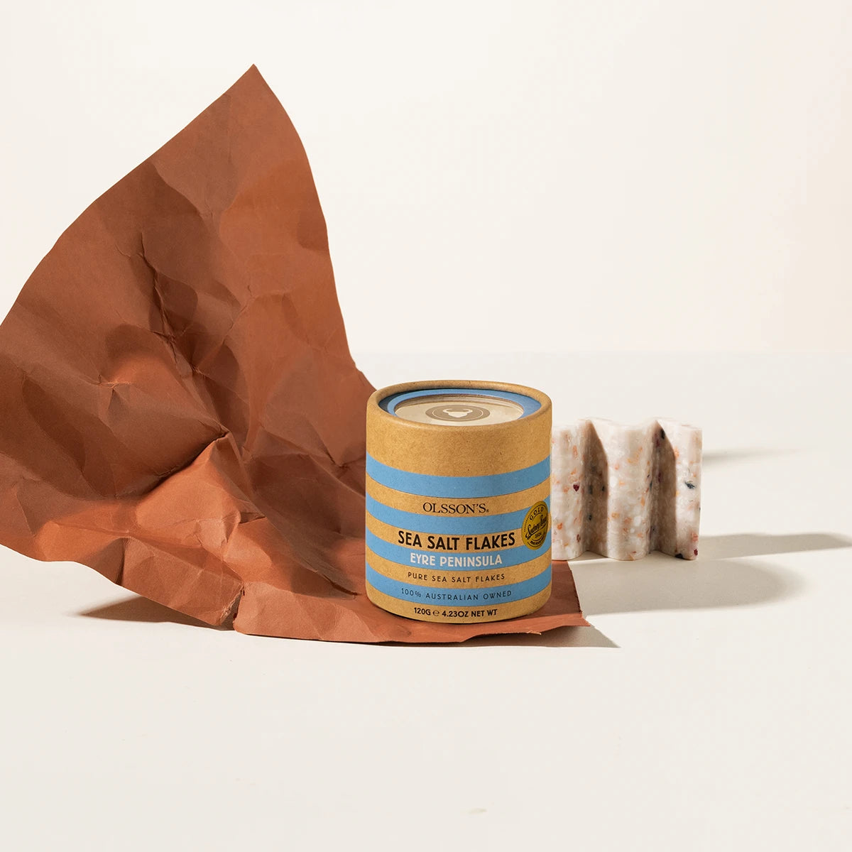 A canister of Olsson's Sea Salt Flakes Blue Flavored Pure Sea Salt Flakes sits on a white surface next to a crumpled brown paper bag and a small block of salt.