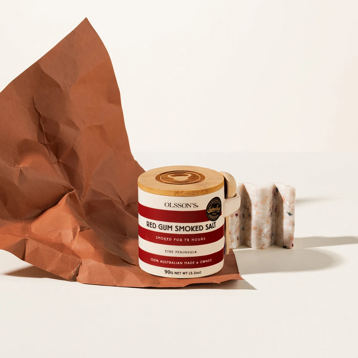 A jar of Olsson's Red Gum Smoked Salt sits on a white surface next to a crumpled brown paper bag and two salt blocks, photographed in a clean studio setting.