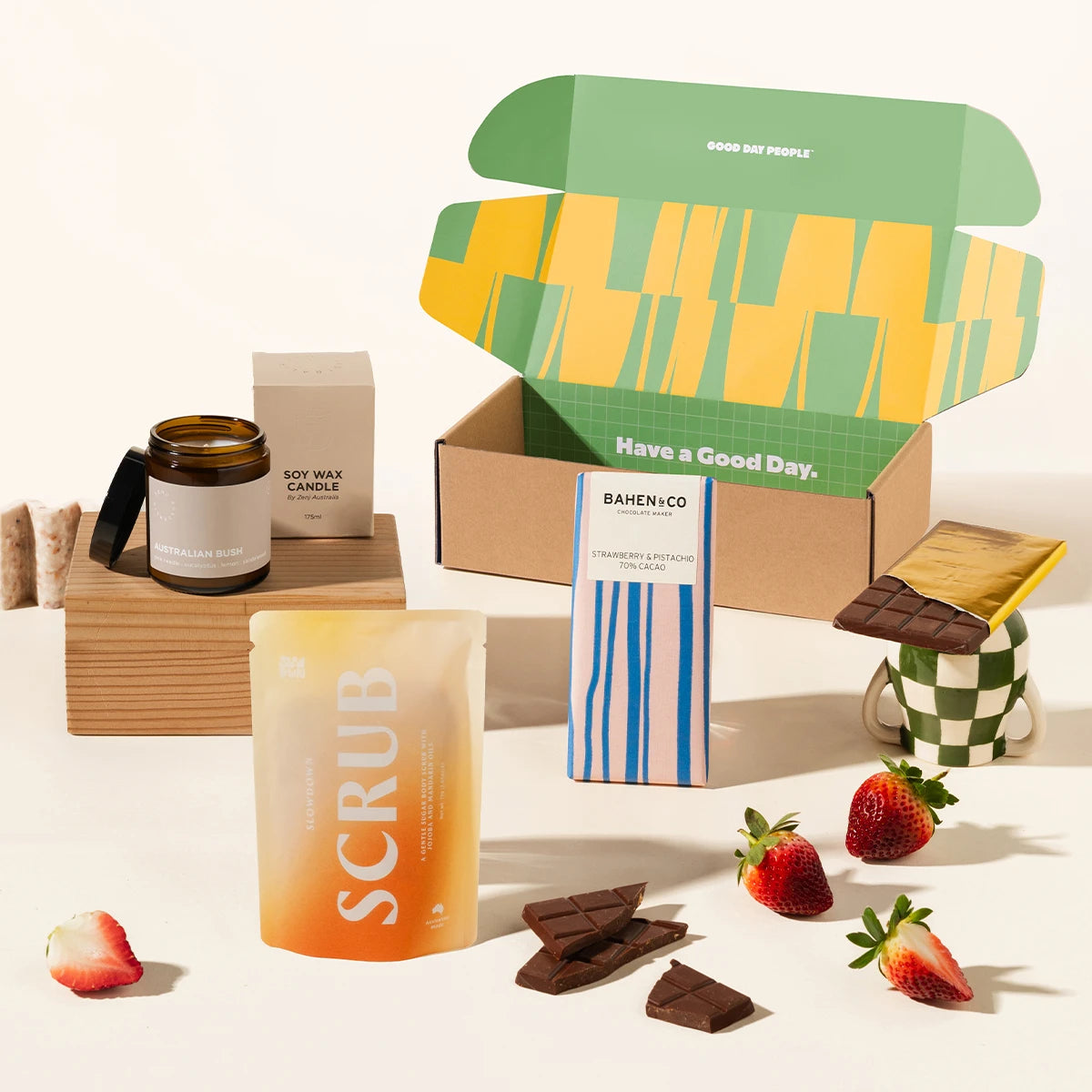 A curated gift set arranged on a clean white surface with an open gift box with gift contents styled around it. Visible items include a scrub pouch, a soy wax candle in a glass jar, a chocolate bar.