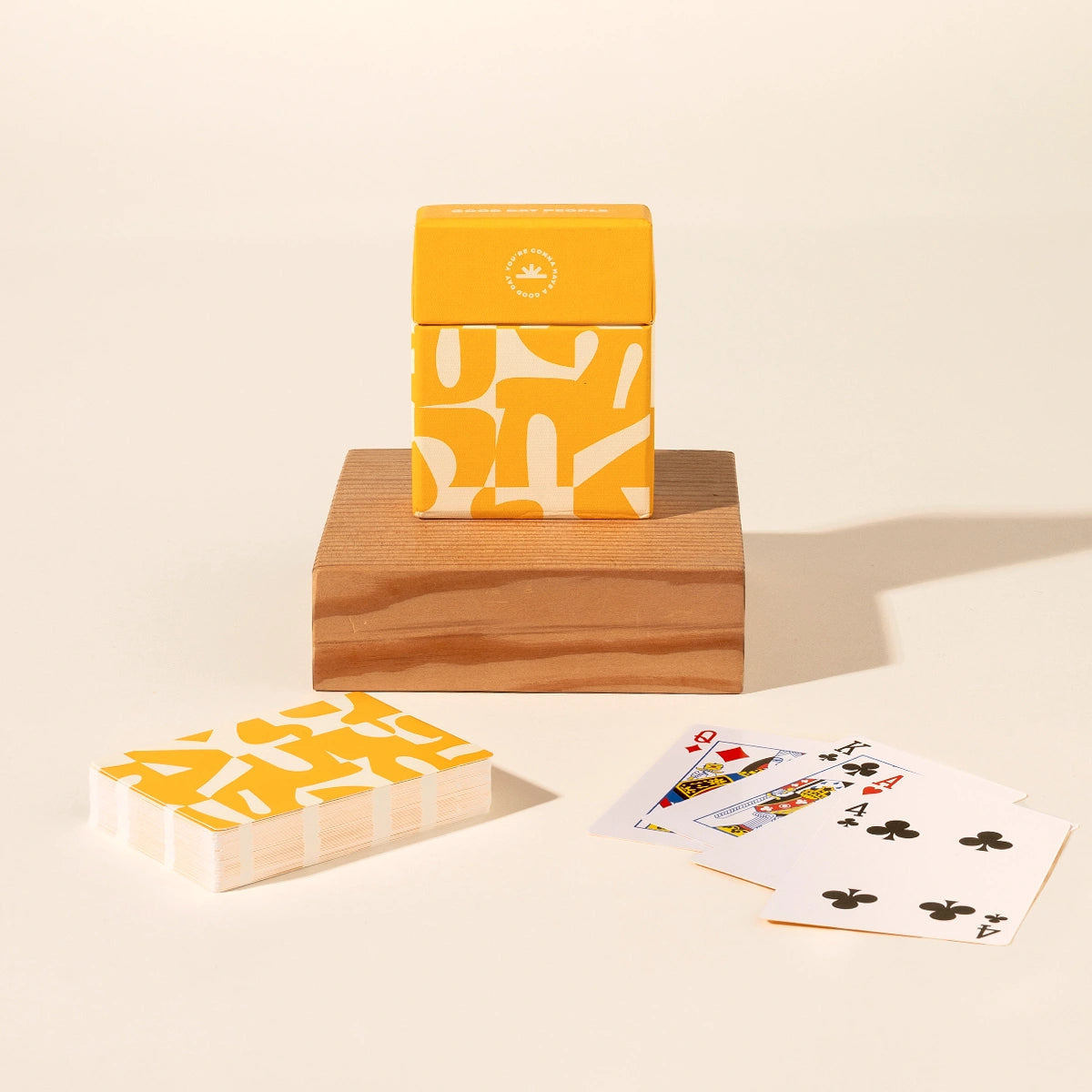 A deck of patterned playing cards with a matching yellow and white box sits on a wooden block, with a second deck and a few fanned cards on a cream background.