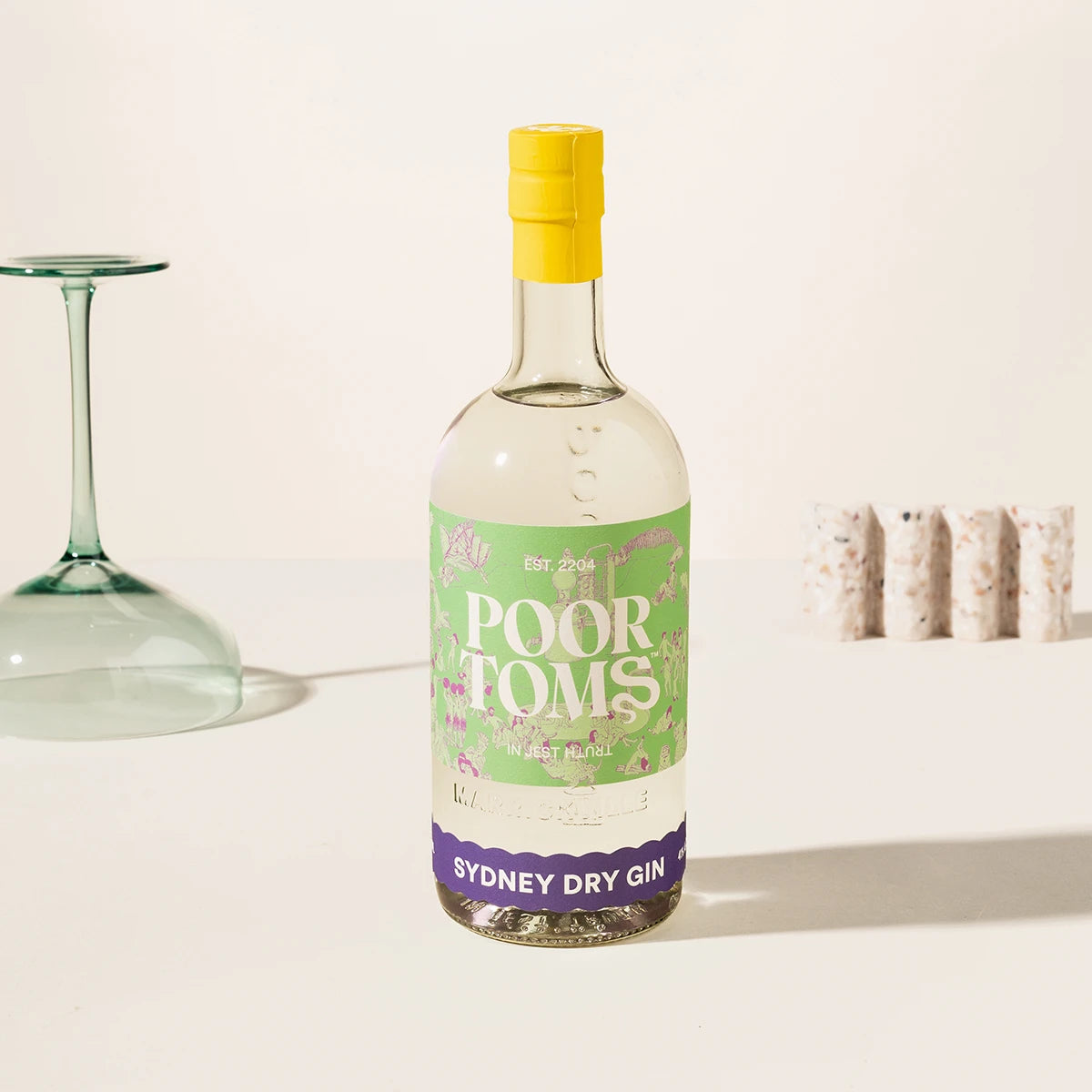 A bottle of Poor Toms Sydney Dry Gin with a yellow cap and green label, set on a white surface with a wine glass and corks in the background.