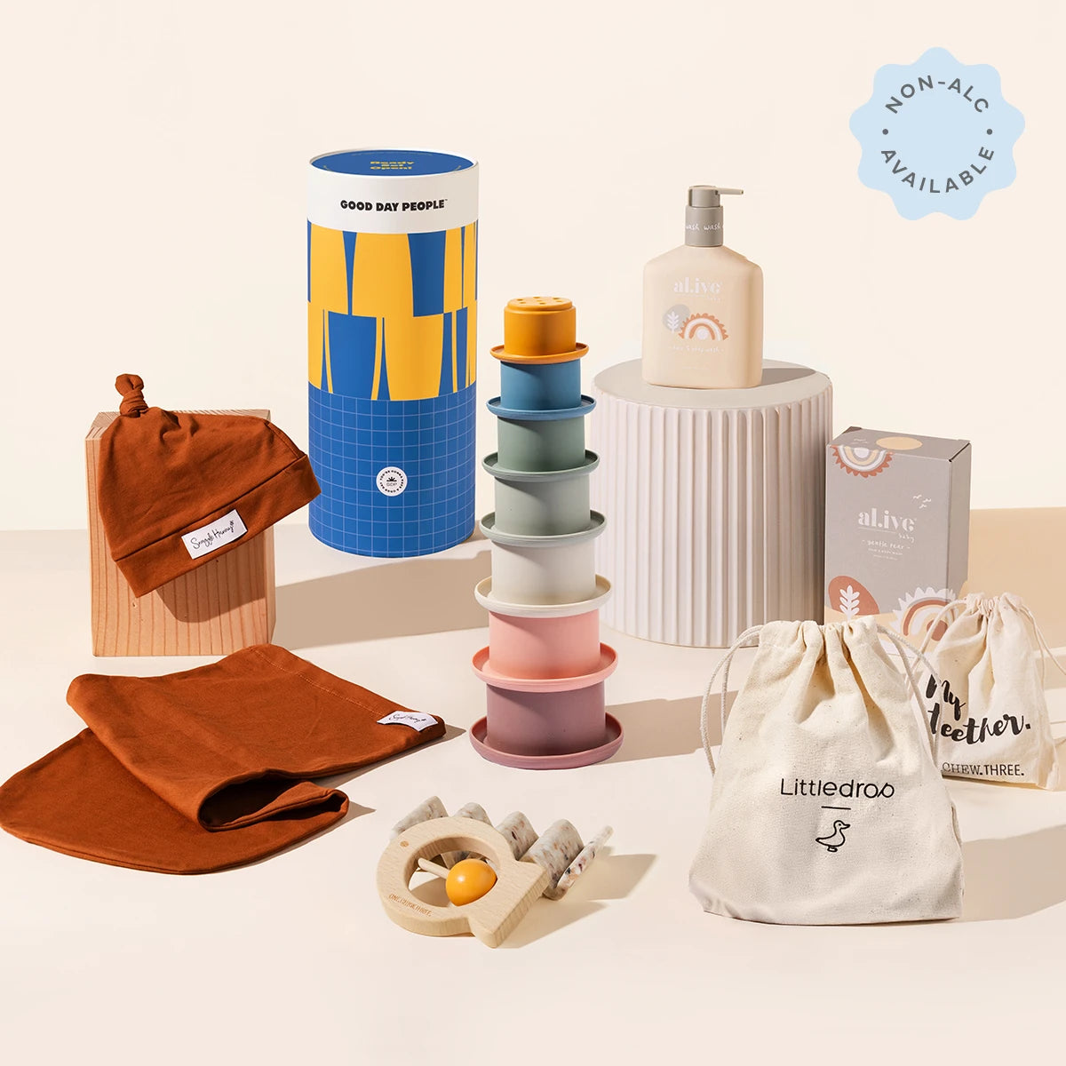 A curated baby gift set including a blue and yellow cylinder, a baby body wash, a stacking toy of pastel silicone cups, a rust-colored jersey swaddle and beanie set, a wooden teething ring.