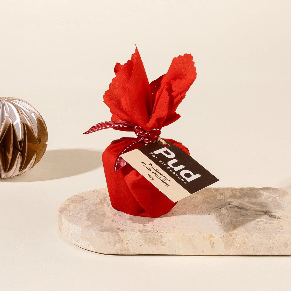 A wrapped mini Christmas pudding in bright red fabric tied with a red ribbon and a brown tag reading 'Pud' styled on a light stone slab against a warm cream background.