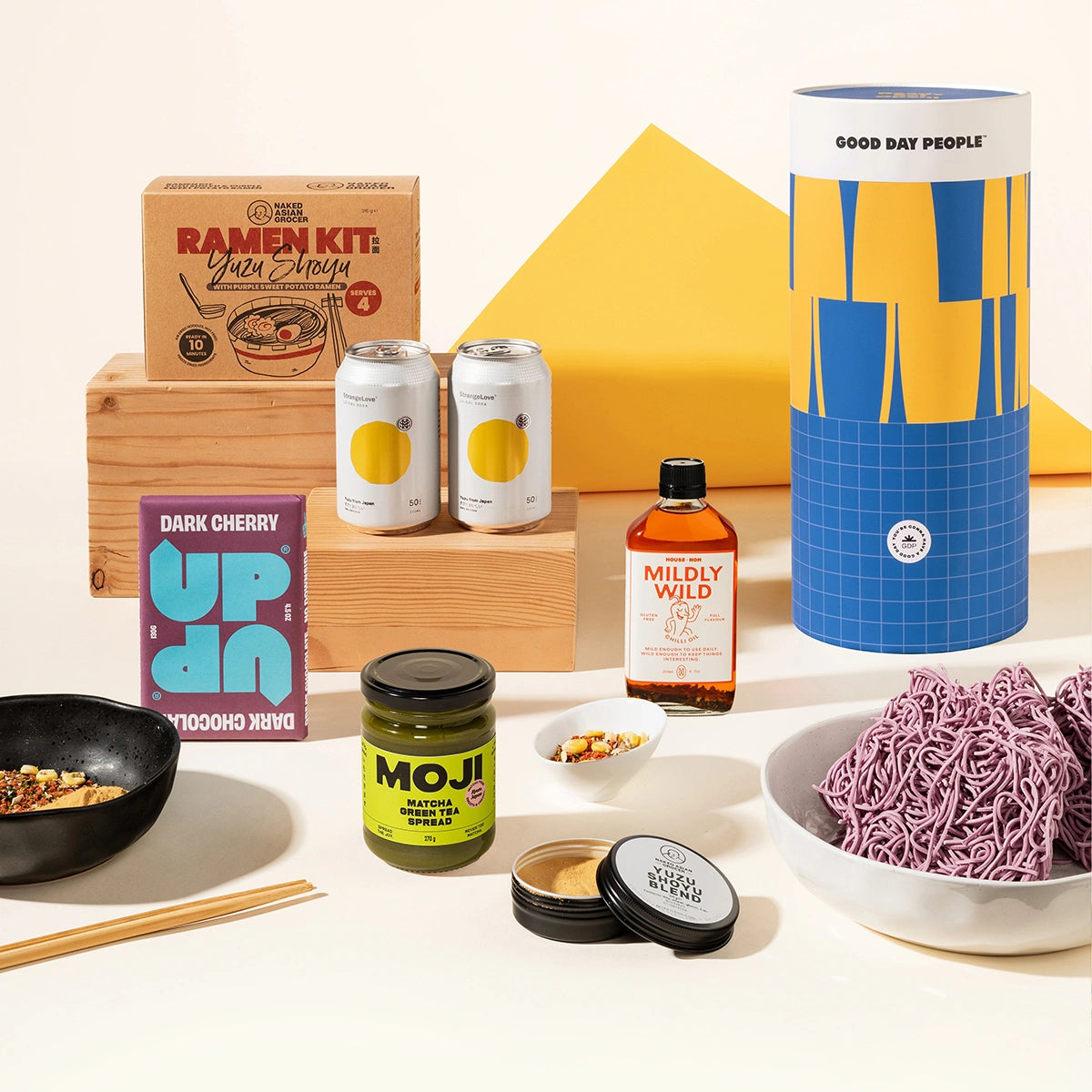 A curated flat-lay of multiple packaged food and beverage products on a warm cream background, including a ramen kit box, two cans, a bottle of Mildly Wild chilli oil, a jar of Moji matcha spread, a bowl of ramen noodles, chopsticks, and a black dish. Ramen Riko Japanese inspired gourmet gift hamper