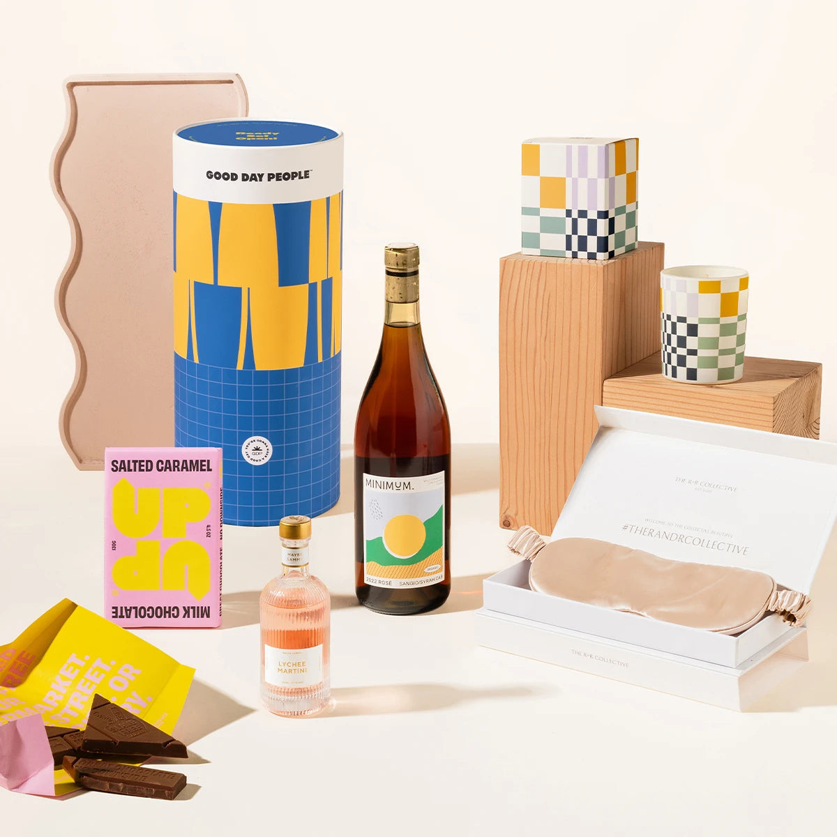 A curated gift set featuring a blue and yellow cylindrer labeled Good Day People, a bottle of rosé wine, a pink salted caramel milk chocolate bar, a small pink bottle containing lychee martini, a blush sleep mask in an open box, and colorful candles and wooden blocks on a clean cream background.