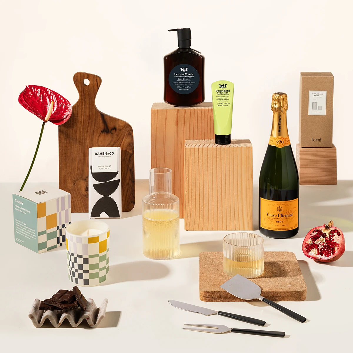 A gift set on a warm off-white background, featuring a bottle of Veuve Champagne, Leif products, a wooden serving board, a Ferm living carafe, chocolate and cheese knives.