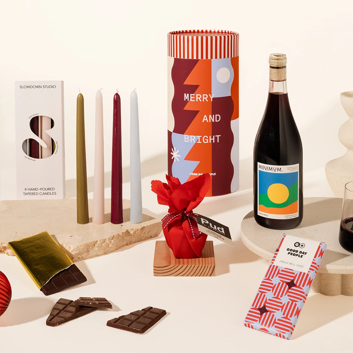 Gift hamper assortment with a bottle of wine, tapered candles (set of 4), a mini christmas pudding wrapepd in red fabric, a block of chocolate in a christmassy red and blue wrapper. Cylindrical packaging with a festive, fun design.