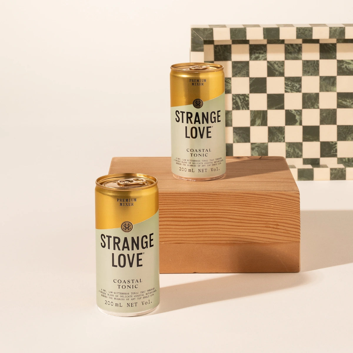 Two cans of Strange Love Coastal Tonic are displayed on a wooden block against a clean white background, with a green and cream checkered box behind them.