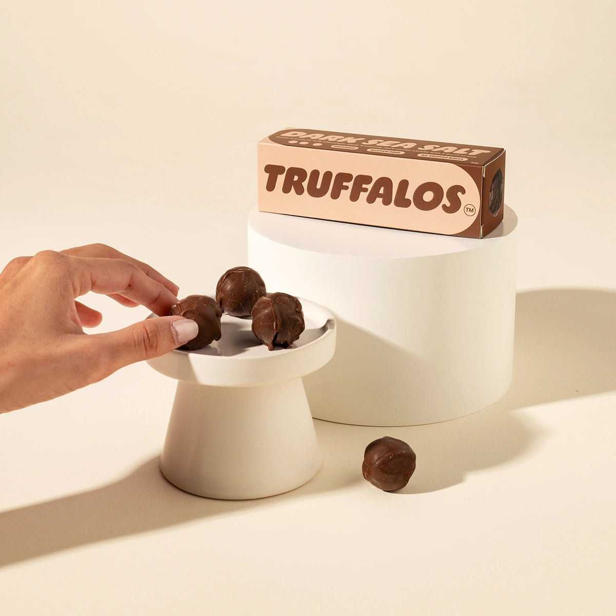 A hand reaches for chocolate truffles displayed on a small pedestal, with a box of Truffalos chocolate truffles standing behind on a larger pedestal against a warm cream background.