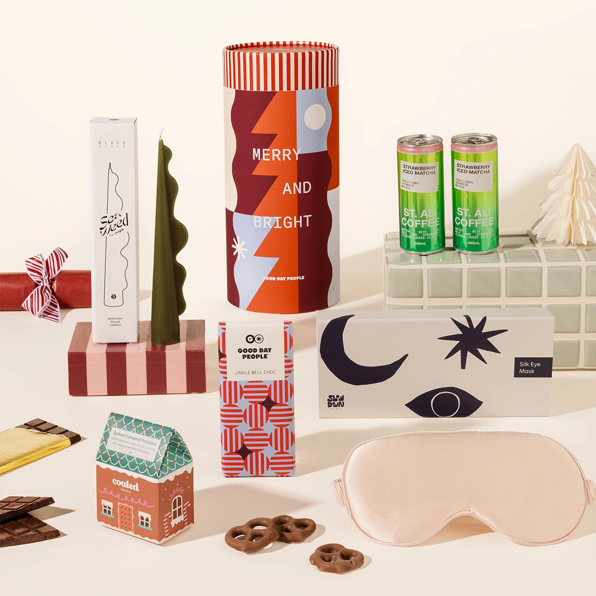 Twinkling Tina Christmas gift box styled with two St Ali Strawberry Iced Matcha cans, a Black Blaze Seaweed Candle, a pink Slowdown Studio silk eye mask, Jingle Bell Choc and chocolate coated pretzels, next to a red and white striped Christmas canister