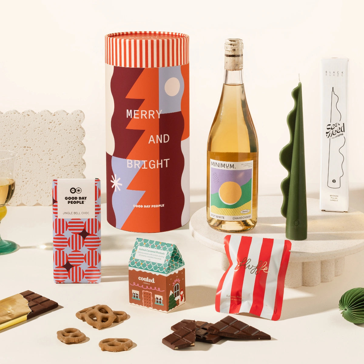 A curated gift set featuring a Christmassy designed canister as the packaging, a bottle of whitewine; a green wavy decorative candle, a block of chocolate with a geometric red and blue design labelled Jingle Bell Choc, a gingerbread house shaped box with pretzels in it and a red and white striped pouch containing fairy floss.