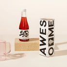 Bottle of red grape, orange and pepperberry non-alcoholic drink with a white label and black text, next to a box labeled 'AWESOME' on a beige background.