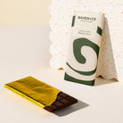 A BAHEN&CO chocolate milk bar in its white and green wrapper, shown next to a partially unwrapped gold foil wrapped chocolate bar on a clean white surface.