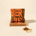 A stand-up pouch of Beer Nuts Jerk Almond Mix sits on a small wooden display block with a few loose nuts placed in front, photographed on a warm off-white studio background.