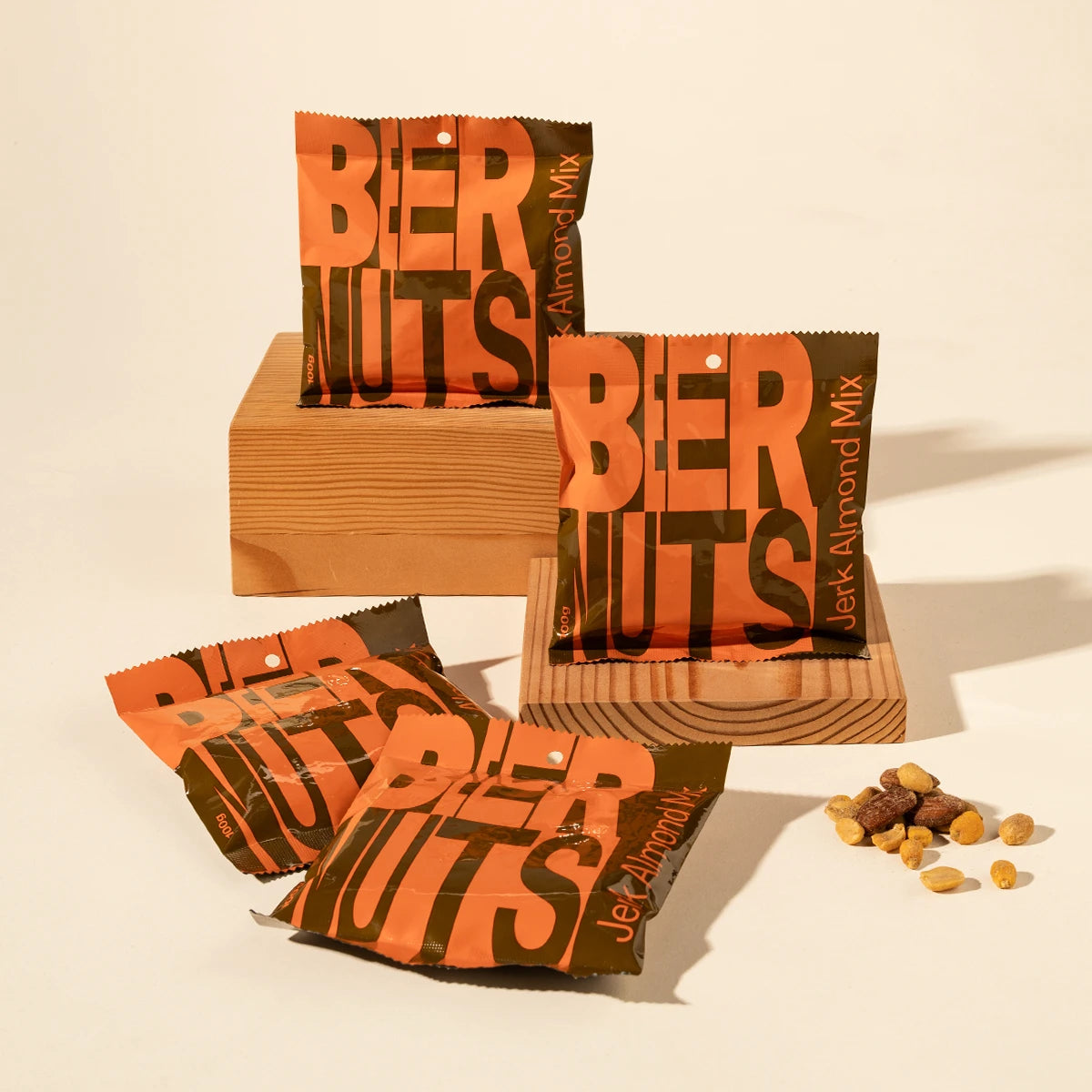 4 bags of Beer Nuts Jerk almond mix are arranged on and around light wood blocks on a warm cream background, with a few nuts scattered nearby. 