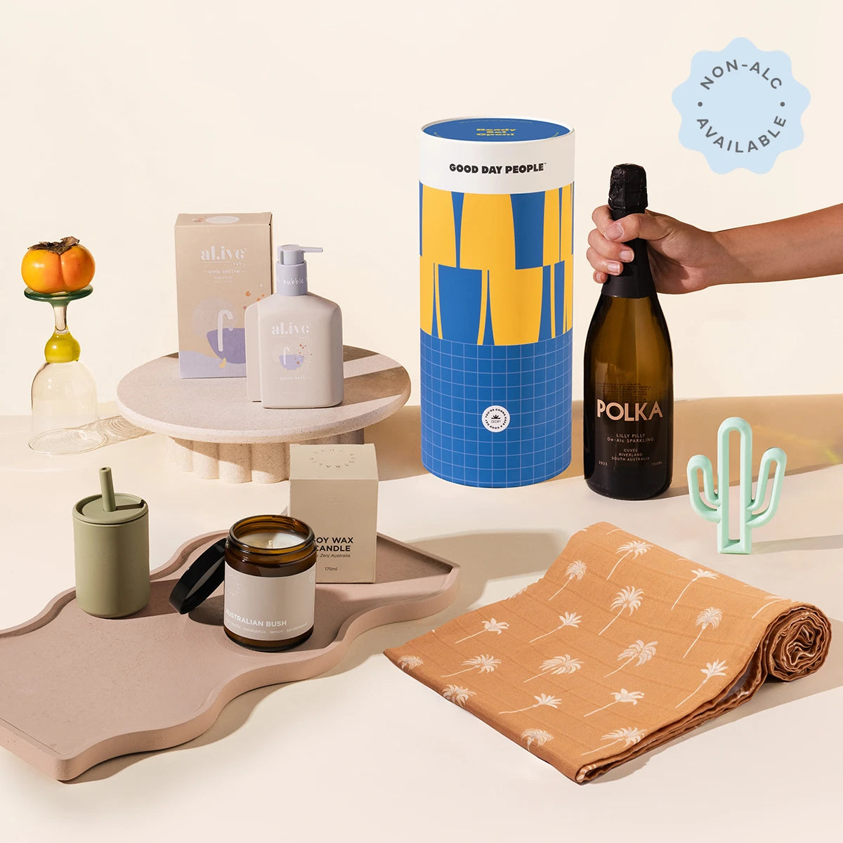 A styled shot featuring a baby hamper. It comed in a stylish blue and yellow cylinder. The hamper contains your drink of choice, a cactus teether, a swaddle, a candle, a silicon sippy cup and baby body wash.