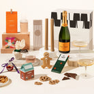 Assorted holiday gift hamper including a bottle of champagne, delicio korean bbq nuts, pillar candles, ferm living carafe and glass set, 2 champagne coupes, chocolate, gingerbread man, salted caramel pretzels and a gin bauble. All packaged in a beautiful gift box. 