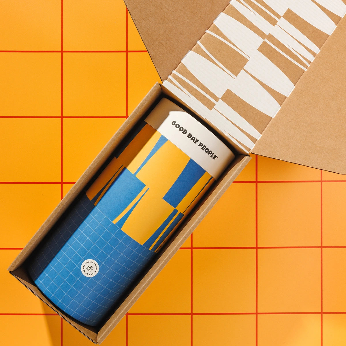 A Good Day People blue and yellow patterned insulated tumbler with a white lid sits inside an open brown cardboard gift box, photographed from above on an orange tiled background.