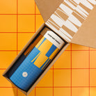 A Good Day People blue and yellow patterned insulated tumbler with a white lid sits inside an open brown cardboard gift box, photographed from above on an orange tiled background.