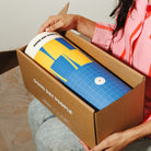 A person holding an open cardboard box while revealing a rolled Good Day People product wrapped in brown paper. The product has a blue and yellow geometric design with a white top section and visible Good Day People branding.