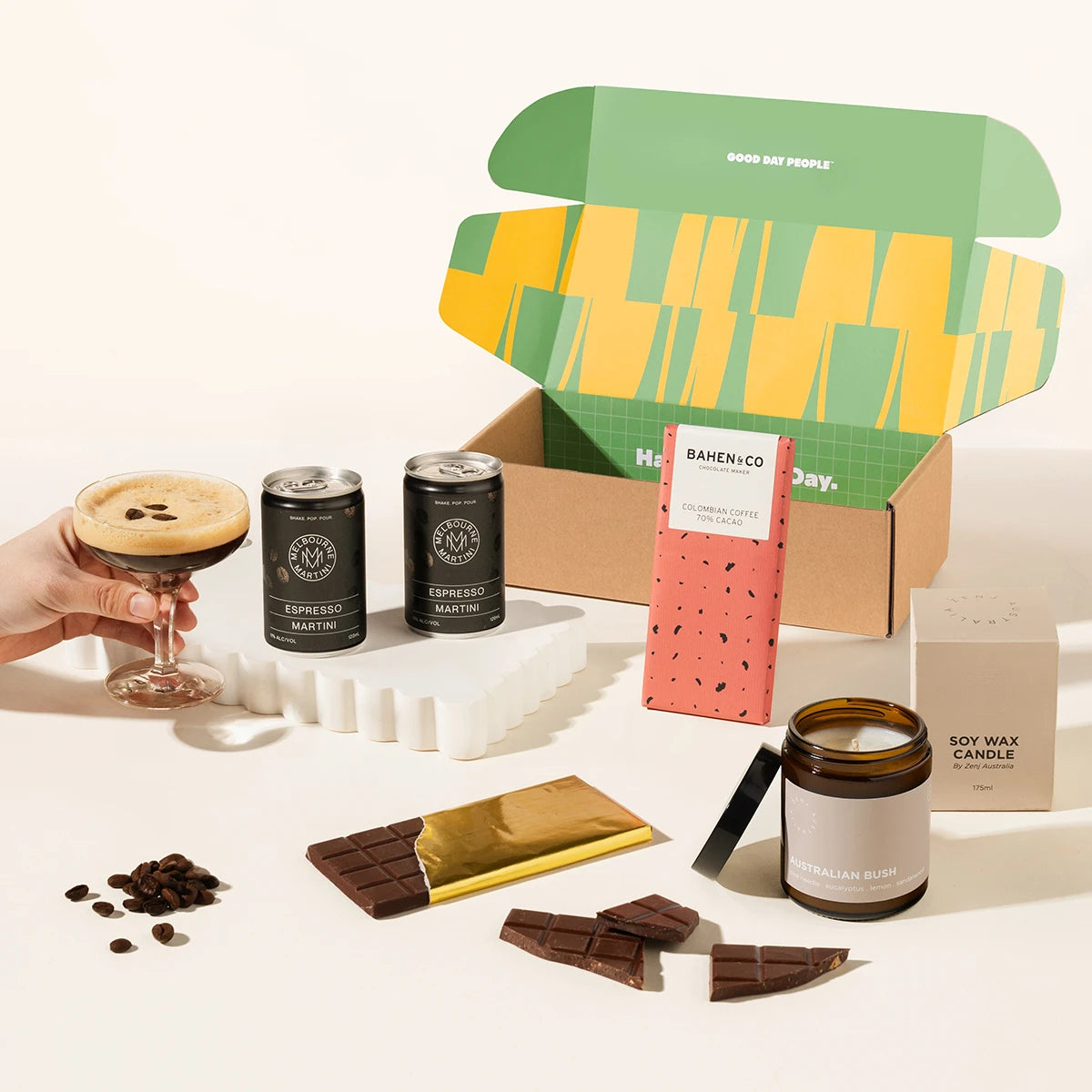 A curated coffee gift set arranged on a warm off-white surface, featuring a patterned gift box, two black espresso martini cans, a hand holding a latte with espresso, a chocolate bar, a soy wax candle jar, and scattered coffee beans. 