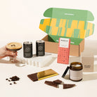 A curated coffee gift set arranged on a warm off-white surface, featuring a patterned gift box, two black espresso martini cans, a hand holding a latte with espresso, a chocolate bar, a soy wax candle jar, and scattered coffee beans. 