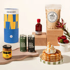 A styled product arrangement on a warm cream background featuring a blue and yellow cylinder,  two ST. ALI cold brew coffee cans, a jar of Koji caramel spread, pancake mix, maple syrup, a stack of pancakes topped with whipped cream and caramel sauce, and a small white cup of strawberries, all arranged on wooden blocks and a wooden board.