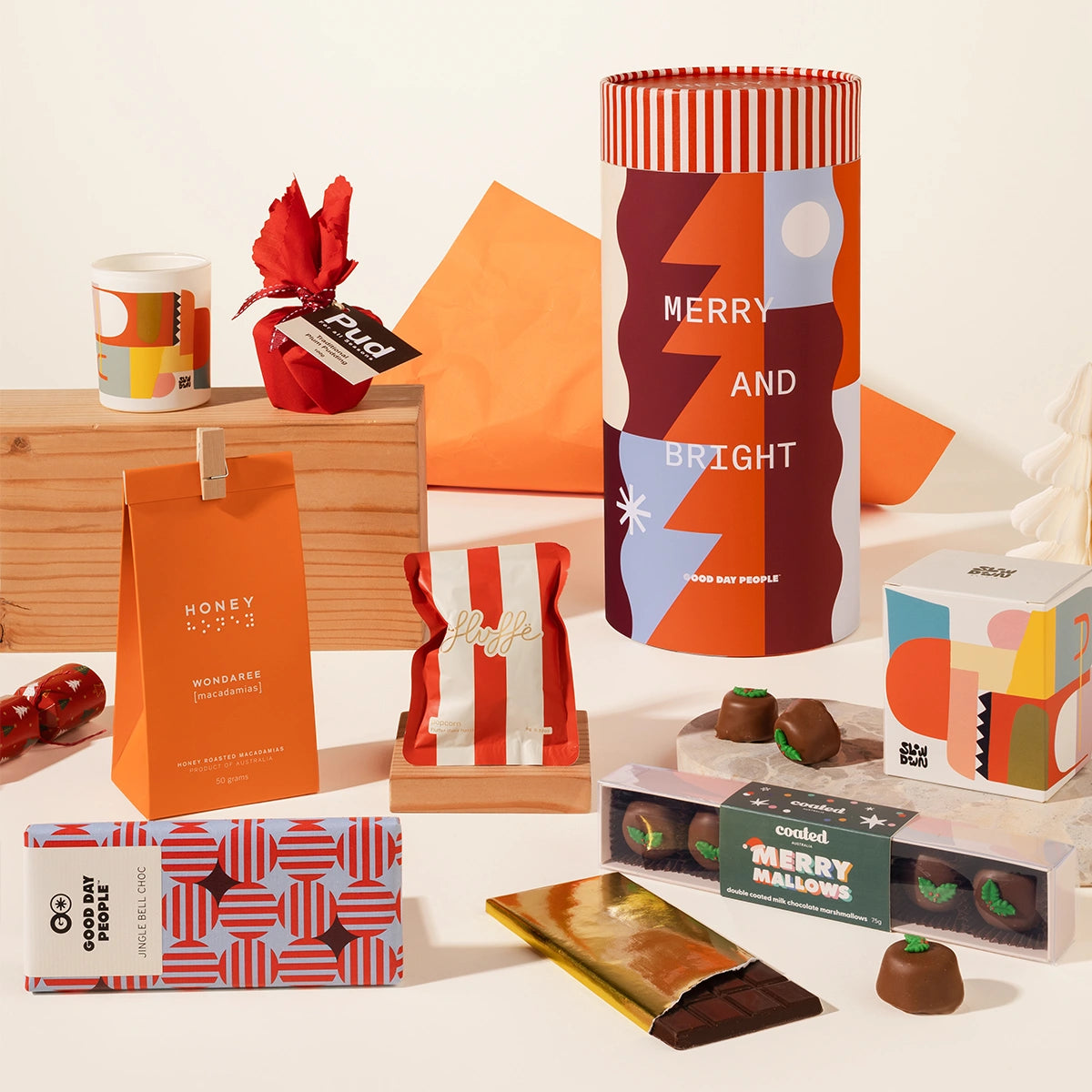 A curated gift set featuring a cylindrical packaging with a colorful geometric pattern and 'MERRY AND BRIGHT' text, a mini christmas pudding wrapped in red fabric,alongside a patterned candle, a wrapped chocolate, a box of macadamias, and assorted chocolates on a clean white background.