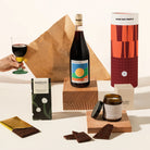 A curated gift set featuring a bottle of wine, a designed Good Day People cylinder, a chocolate bar, a jar of soy wax candle.