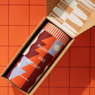 A cylindrical insulated tumbler with a festive geometric design in orange, burgundy, light blue, and white, presented inside an open kraft paper gift box. The tumbler features the text 'MERRY AND BRIGHT' and 'GOOD DAY PROUDLE'.