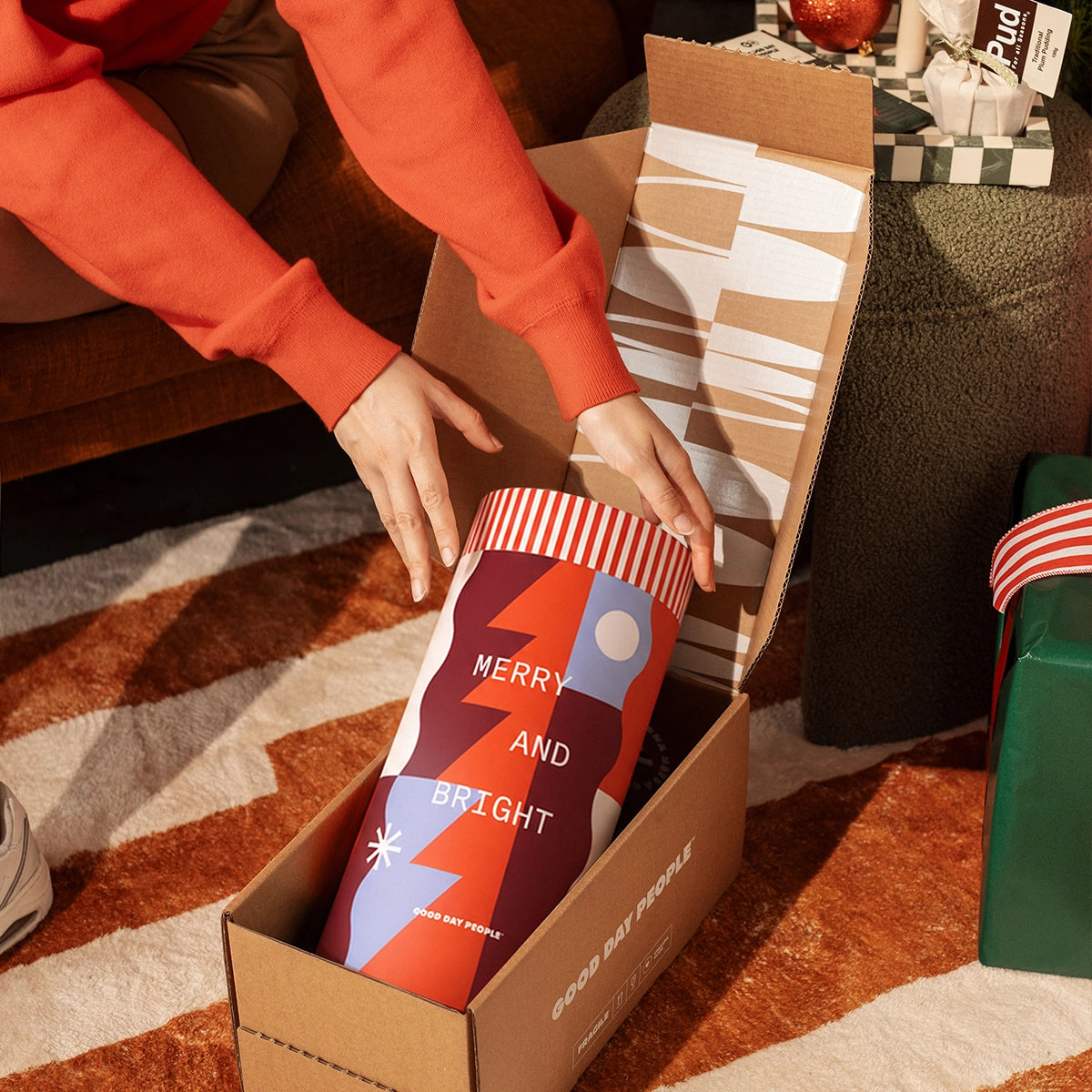 A person in an orange sweatshirt opens a cardboard gift box containing a cylindrical tumbler with a festive holiday design. The tumbler features the text 'MERRY AND BRIGHT' over abstract geometric shapes in red, orange, purple, and white.
