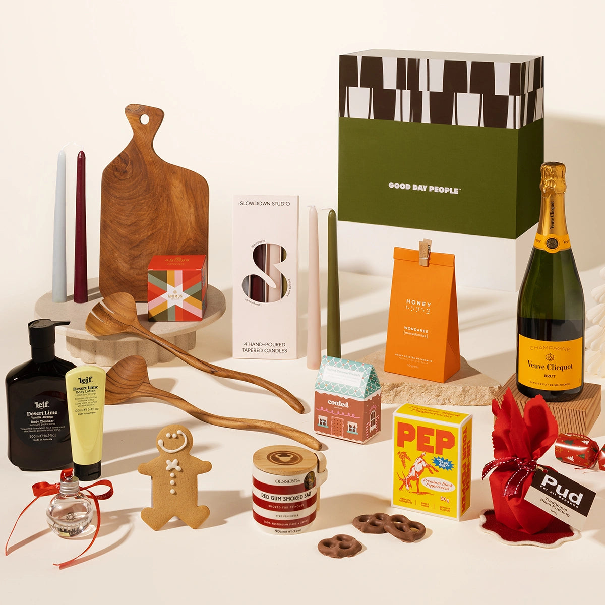A curated gift basket arrangement on a warm cream background, featuring a green and black Good Day People box, a bottle of Veuve Clicquot champagne, a yellow Pep peper box, gingerbread cookies, candles, wooden spoons and cutting board, Leif body wash and soap, a gin bauble, pretzels, gourmet salt, christmas pudding