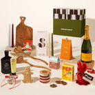 A curated gift basket arrangement on a warm cream background, featuring a green and black Good Day People box, a bottle of Veuve Clicquot champagne, a yellow Pep peper box, gingerbread cookies, candles, wooden spoons and cutting board, Leif body wash and soap, a gin bauble, pretzels, gourmet salt, christmas pudding