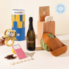 Delivered Diana New Parent & Baby Gift Hamper. A curated gift set featuring a bottle of Polka sparkling wine, a blue and yellow canister, a small plush lion toy, a pink patterned chocolate bar, a small wooden stand with a konjac sponge, and a brown silicone bib with a watermelon cutout, arranged on a light beige surface.
