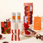A curated gift assortment featuring various packaged food products, including a large cylindrical gift packaging canister with a Christmas design, macadamias, fairy floss, 2 x strangelove sodas, mount zero olives and a mini christmas pudding.