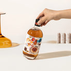 A hand holds a clear glass bottle filled with Christmas Gin by Four Pillars against a clean white background. The bottle has a colorful illustrated label and a black cap.
