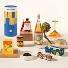 A curated gift-style product arrangement on a light neutral background, including a blue and yellow cylindrical container labeled GOOD DAY PEOPLE, a bottle of wine, a jar of Koji salted caramel, a yellow PEP box, a boxed snack bar labeled ROCKY ROAD.