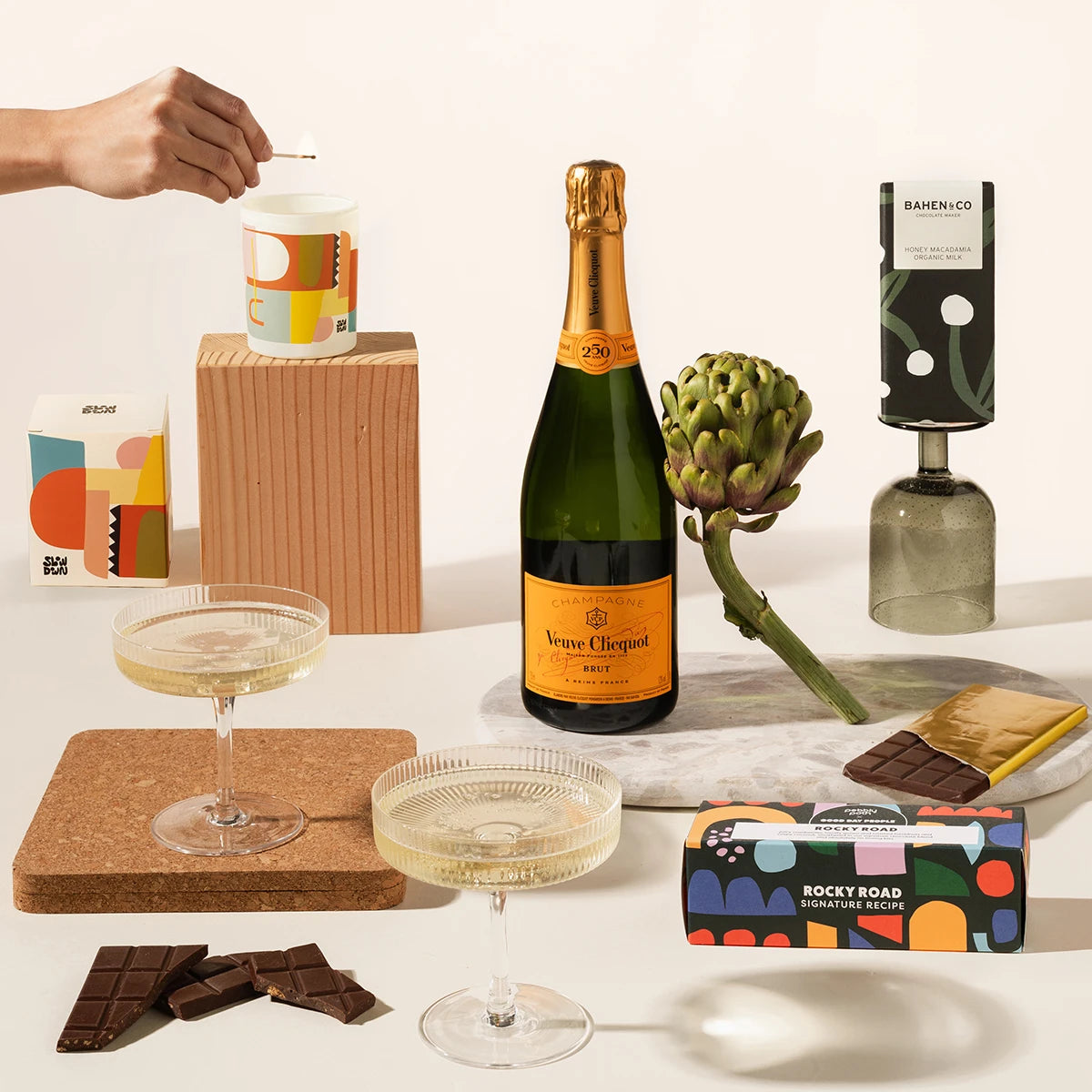 A styled gift set featuring a bottle of Veuve Clicquot champagne, a box of Rocky Road chocolate, a candle, and a pair of Ferm Living champagne glasses.